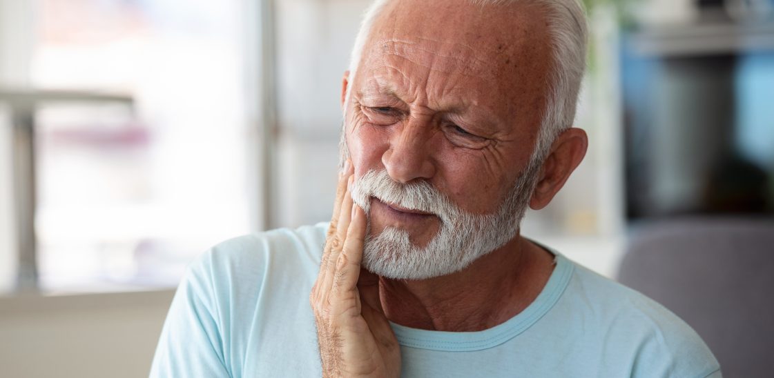 older man with grey beard winces in pain with his hand to his face