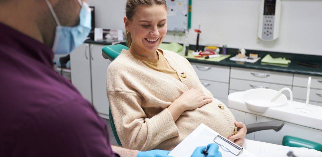 Dentist hygienist prescribing a dental prophylaxis treatment to a young pregnant woman, smiling while sitting on dentist's chair. The concept of a preventive examination by a dentist during pregnancy