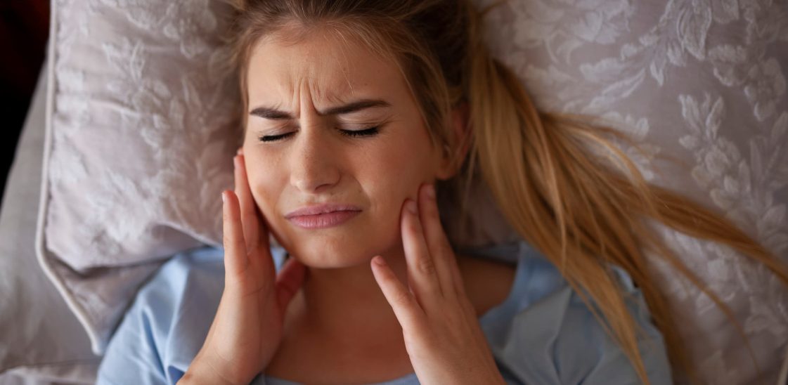 A woman holds her jaw in pain while lying in bed.